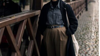 A person wearing dark academia fashion with vintage blazer, turtleneck, and classic book in hand, standing near an old library